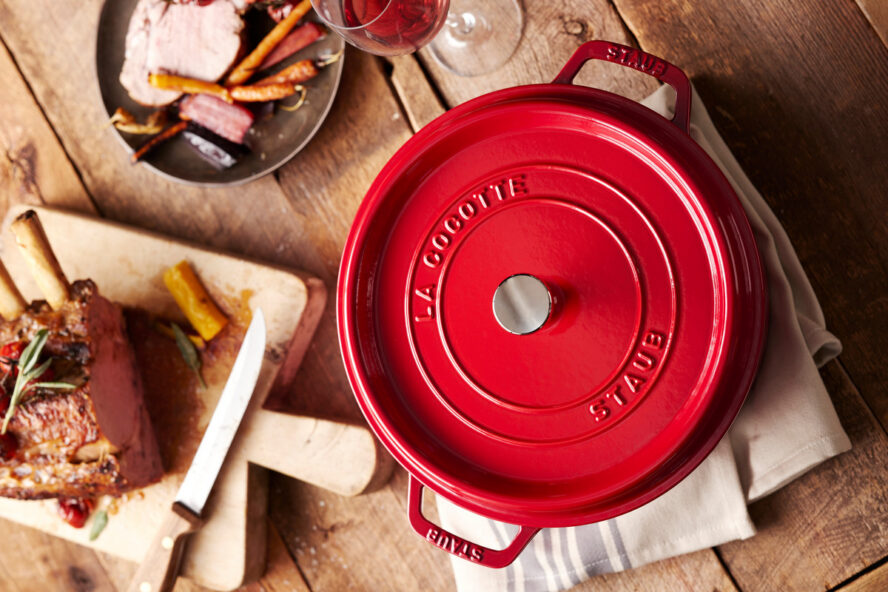 Image of a cherry red Staub round Dutch oven shown on a rustic wooden table next to a rack of lamb.