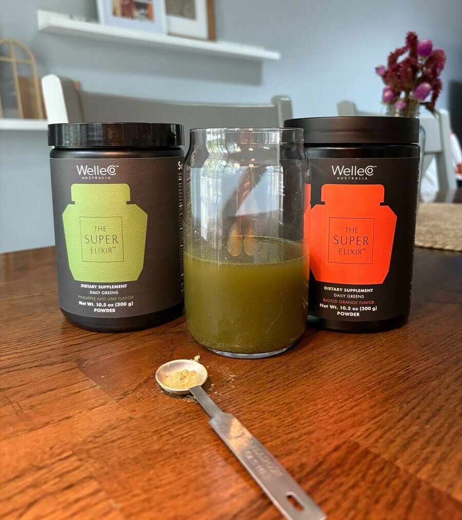 Image of two sleek tubs of WelleCo The Super Elixir in the flavors Blood Orange and Pineapple & Lime, shown with a glass of greens powder and a small spoon in between them.