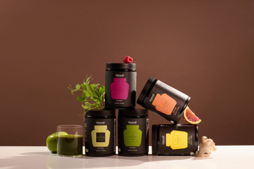 Image of several tubs of WelleCo The Super Elixir in various flavors stacked on top of each other, set against a brown background.