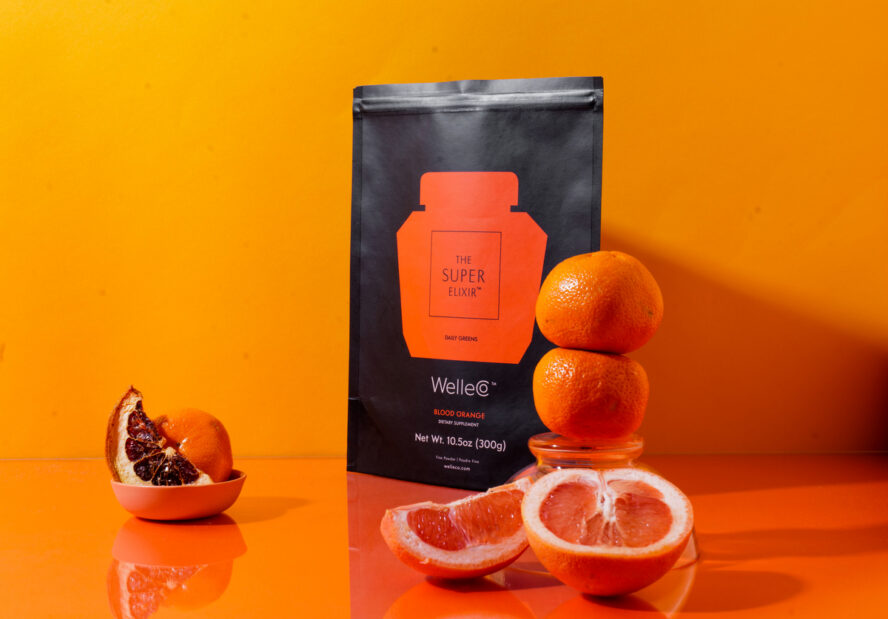 Image of a WelleCo The Super Elixir flatpack in the Blood Orange flavor, set against a vibrant orange background with citrus fruits in the foreground.