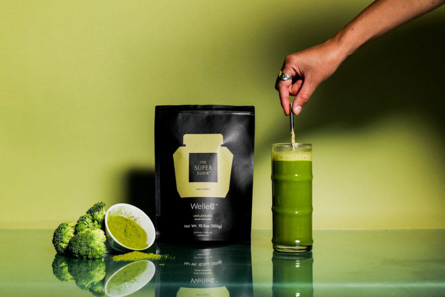 Image of a packet of Unflavoured The Super Elixir from WelleCo, set against a green background with a hand stirring greens powder into a tall glass of water.