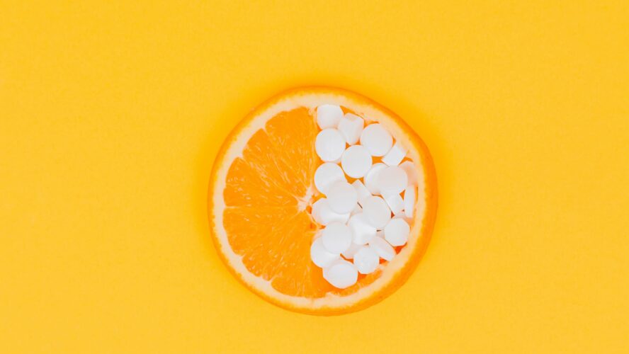 Image of a slice of orange laid flat, with half of the fruit replaced with small white supplement pills.