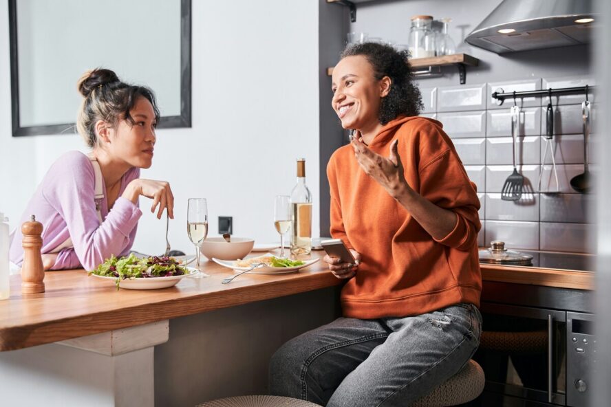 Image of two women sitting together over a kitchen bar, sharing a glass of wine and plates of salad made up of high-fiber foods.