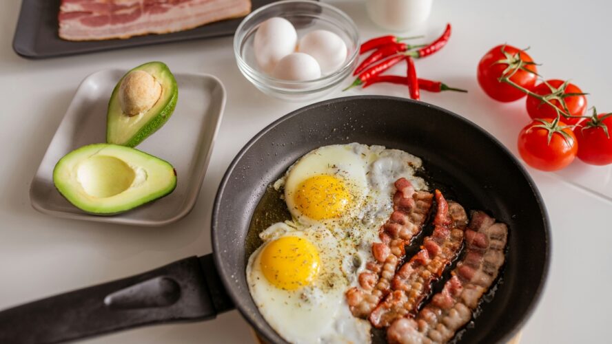 Image of a frying pan with two fried eggs and bacon, ona. counter next to an avocado and tomatoes on the vine.