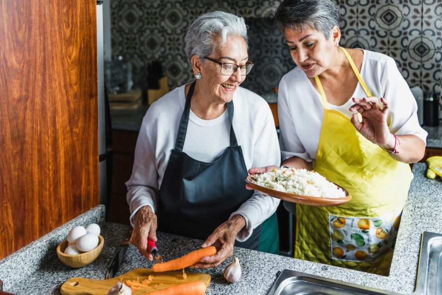 Image of two older women in the kitchen, wearing aprons and smiling as they prepare food together.