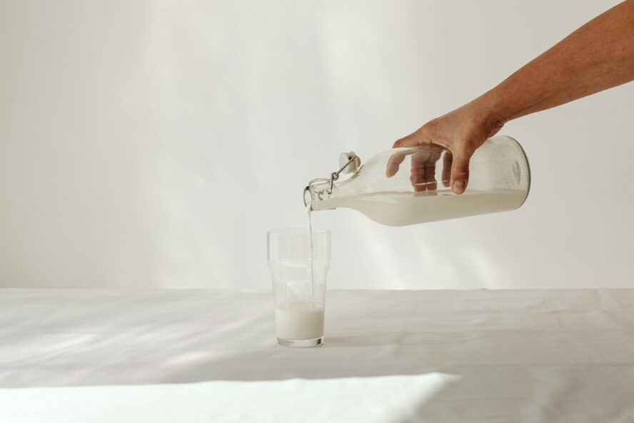 Image of a hand pouring milk from a glass bottle into a glass, all against a white background.