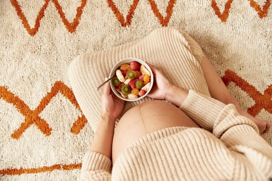 Image of a pregnant woman sitting on a rug on her knees, with a bowl of high-fiber fruits resting in her lap.