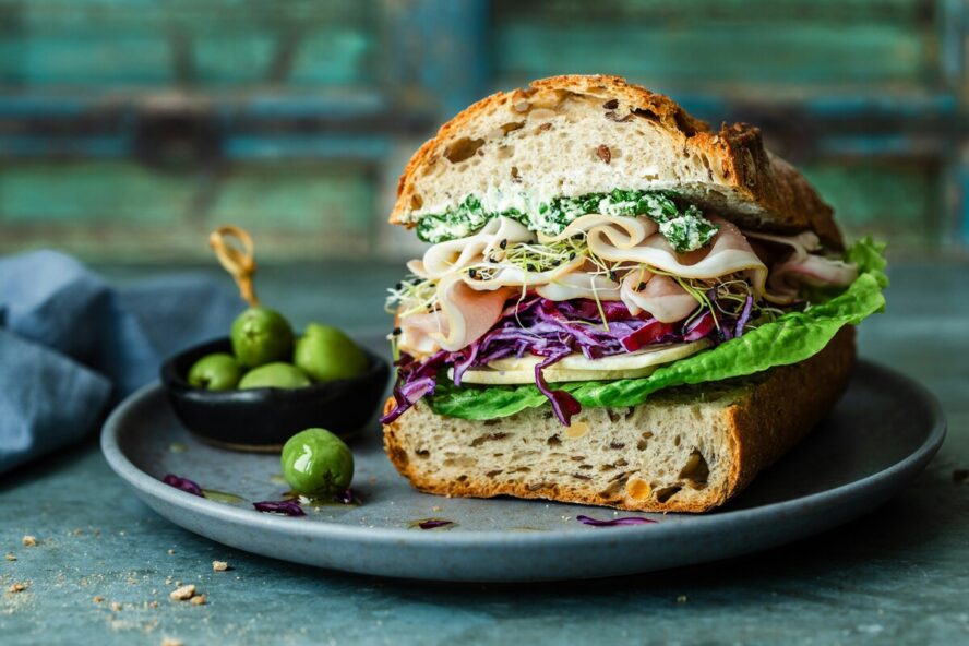 Image of half of a sandwich on a plate made with a hearty whole grain seeded bread, deli meat, and various greens.