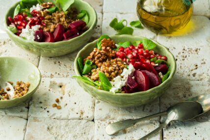Image of a couple of green bowls filled with vegetables and grains, including beetroot, greens, walnuts, and pomegranate seeds.