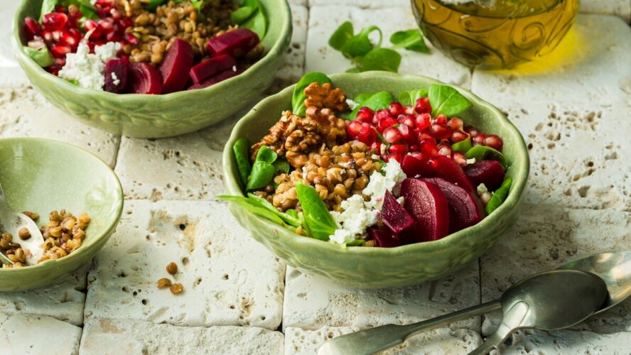 Image of a couple of green bowls filled with vegetables and grains, including beetroot, greens, walnuts, and pomegranate seeds.