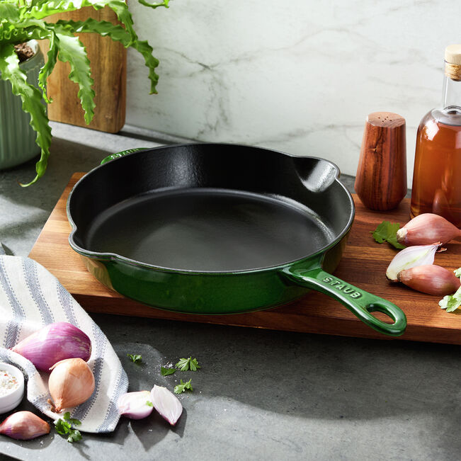 Image of the Staub Traditional Deep Skillet in Basil, sitting on a wooden cutting board next to some shallots and a dishcloth.
