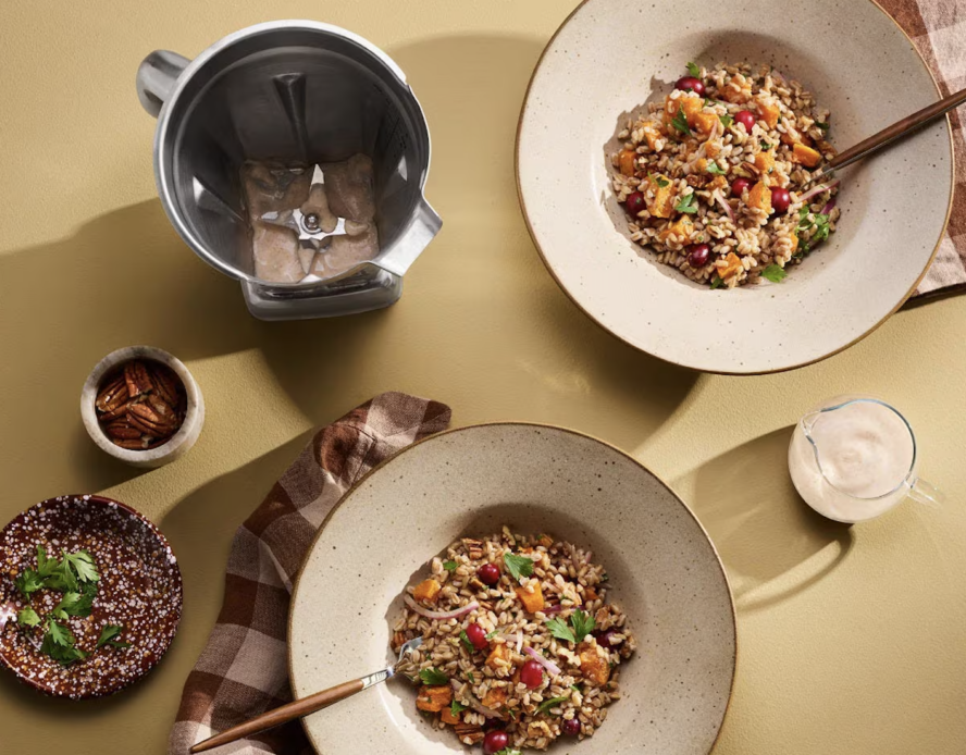 Image of a Vitamix blender with the stainless steel blender jar, shown next to two plates of a fresh barley salad with a blended dressing in a jar.