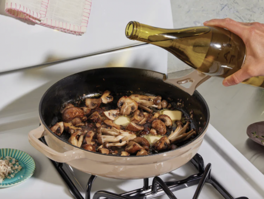 Image of the Our Place Cast Iron Always Pan in the color Steam on a stovetop, with a hand pouring white wine over mushrooms in the pan.