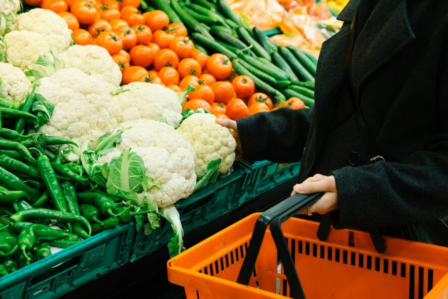 Image of a woman in a black jacket shown from the neck down, standing in the produce aisle of a grocery store holding an orange shopping basket and assessing a head of cauliflower.