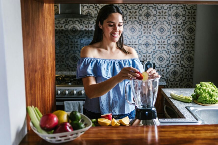 Image of woman putting fruits into a blender in a brightly lit kitchen.