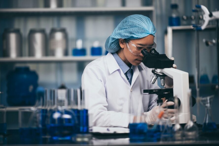 Image of a young woman wearing a hairnet and safety goggles looking at something under a microscope in a lab.