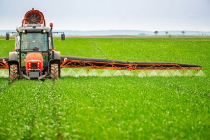 Image of a large tractor driving over a field of crops, spraying herbicides and pesticides as it goes.