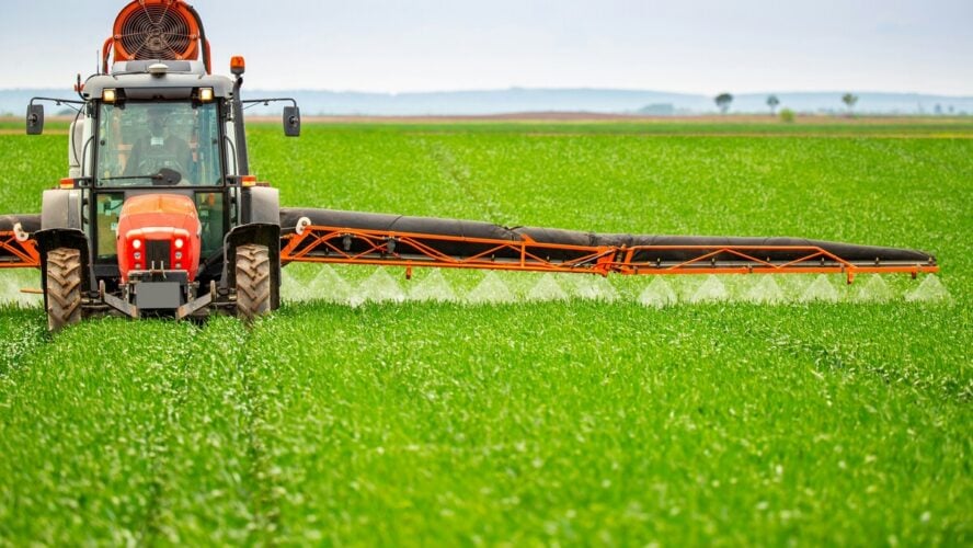 Image of a large tractor driving over a field of crops, spraying herbicides and pesticides as it goes.