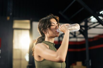 Image of a young woman in a ponytail drinking from a plastic water bottle at a gym.