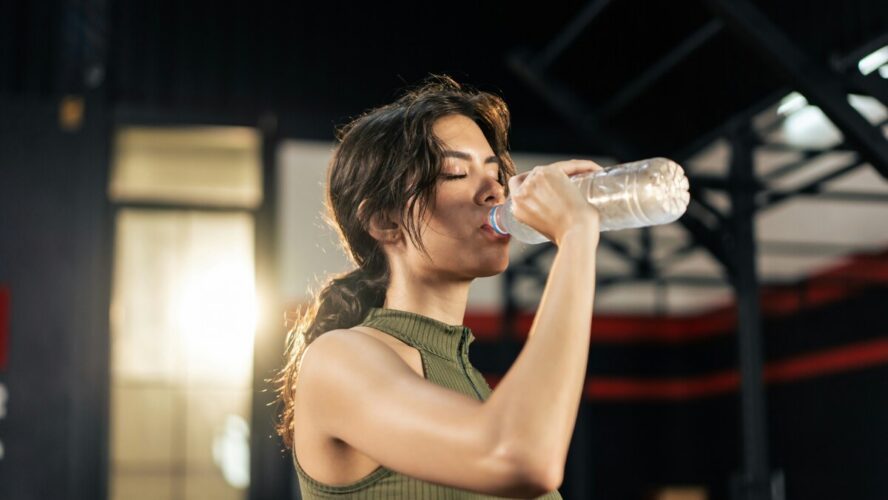 Image of a young woman in a ponytail drinking from a plastic water bottle at a gym.