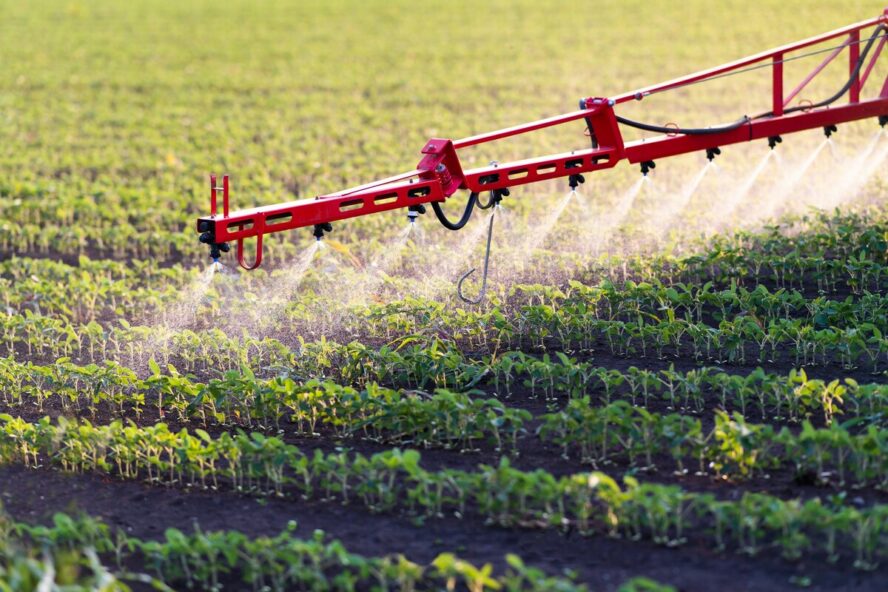 Image of a long spraying extension of a tractor, with a number of nozzles spraying toxic pesticides over a field of crops.