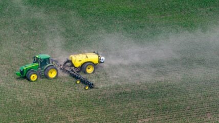 Overhead image of a yellow tractor spraying a glyphosate-based herbicide over a field of crops, with a cloud of smoke billowing around it.