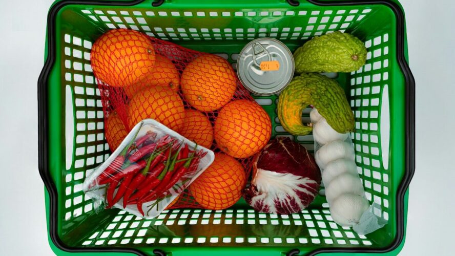 Image of a green grocery basket filled with produce items like oranges, garlic, cabbage, and chilis.