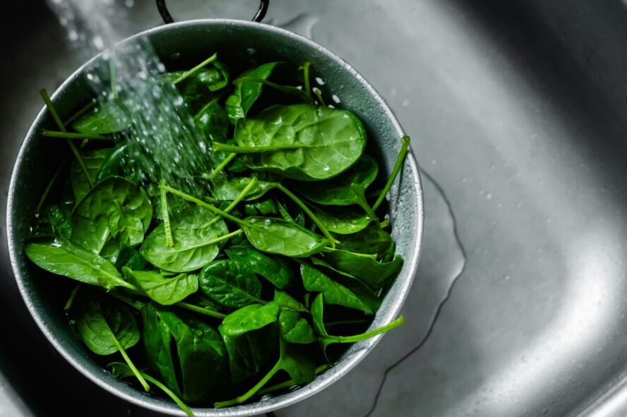 Image of a large silver bowl filled with spinach, sitting under a tap being rinsed with water.