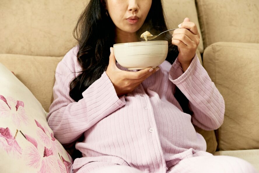Image of a pregnant woman sitting on a couch and eating from a bowl of oatmeal.