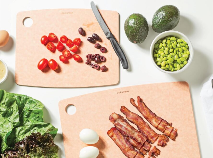 Image of two Epicurean composite cutting boards, one being used to chop olives and cherry tomatoes and the other slicing bacon.