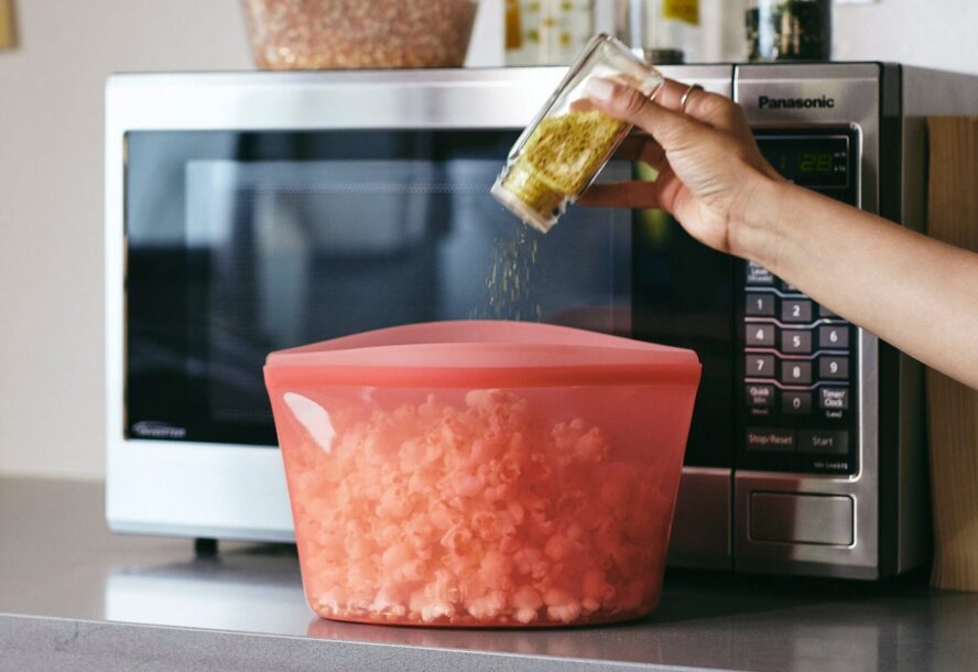 Image of a large Stasher silicone bag being used for microwave popcorn, with a hand seasoning it with nutritional yeast.
