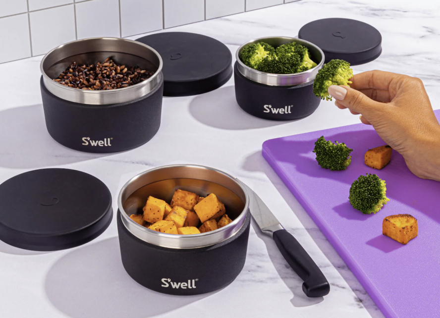 Image of various S'well stainless steel containers open to reveal different roasted and raw vegetables on a kitchen countertop next to a purple cutting board.
