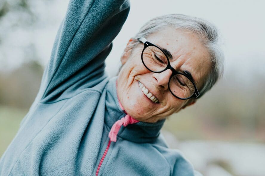 Image of an older woman wearing glasses and stretching with her arm over her head and her eyes closed.