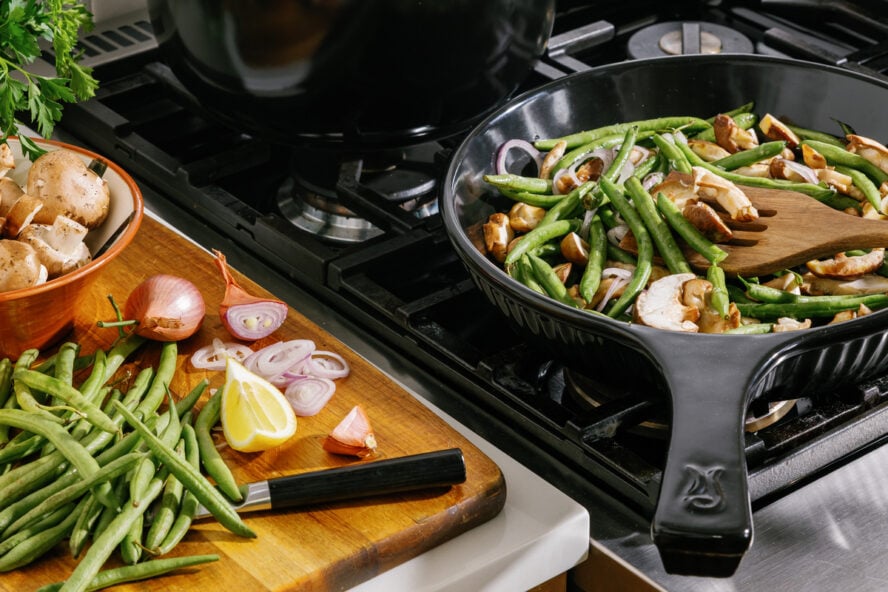 Image of a large Xtrema pure ceramic skillet shown on a stovetop full of green beans and other vegetables, with a cutting board shown on the side.