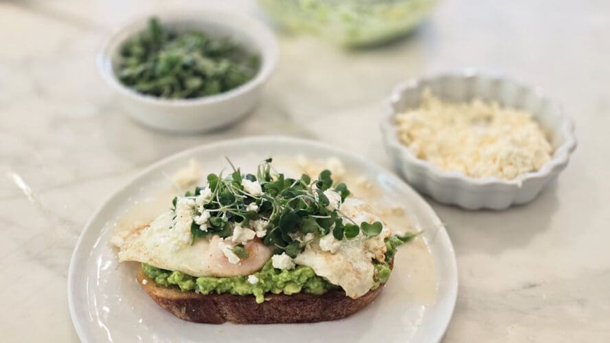 Image of a slice of toast topped with avocado and eggs, with small dishes of microgreens and feta behind the plate.