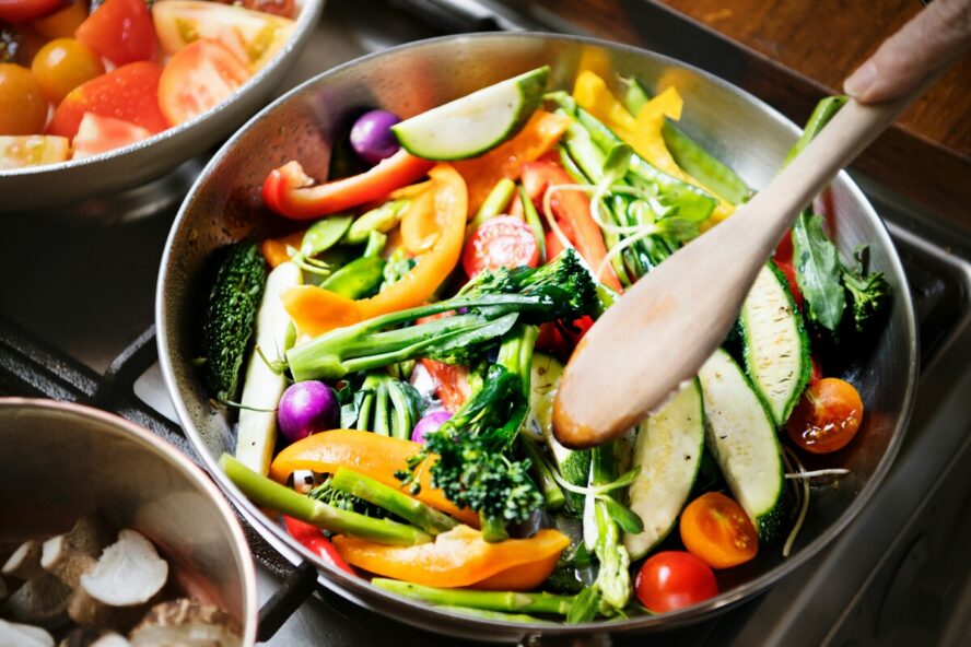 Image of a frying pan on a stovetop full of chopped vegetables being stir-fried with a wooden spoon.
