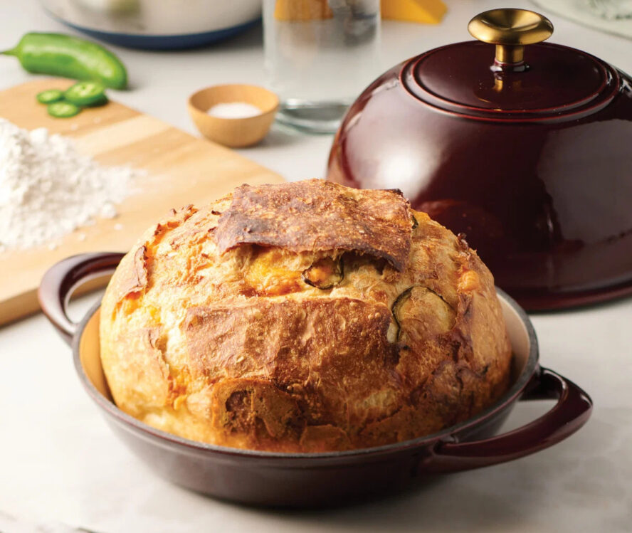 Image of a Tramontina Cast Iron Bread Oven in the color Cranberry, shown with a large loaf of jalapeño bread.