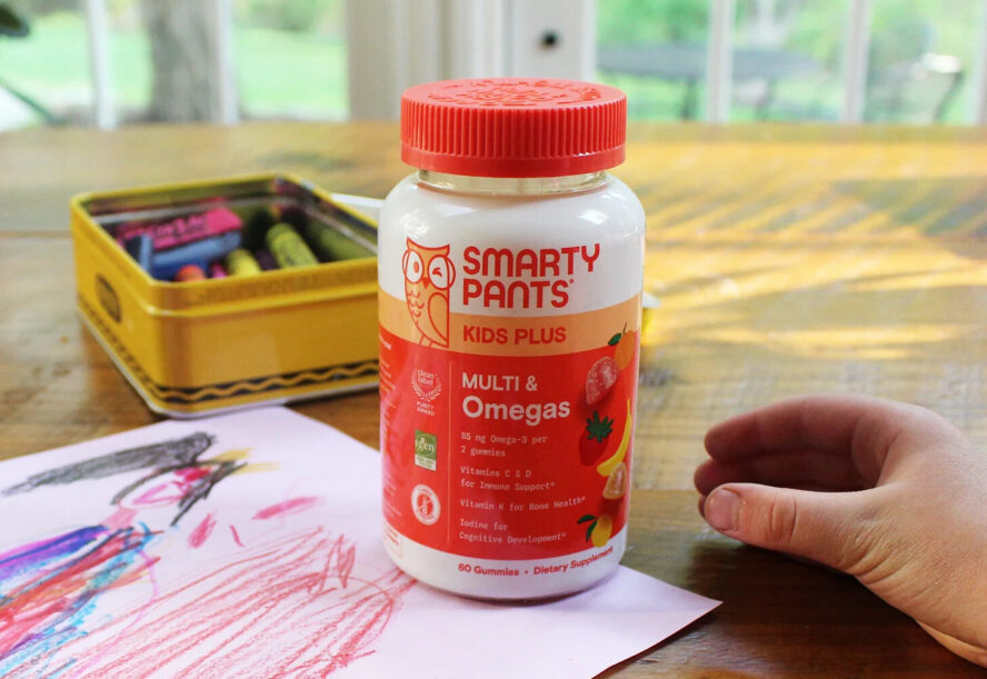 Image of a bottle of SmartyPants Multi & Omegas vitamins, shown on a table with kids' arts and crafts.