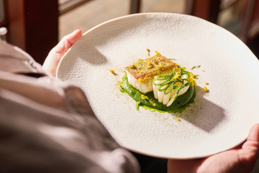 Image of a beautifully plated dish of food, with a pair of hands carrying it to serve it to a guest.