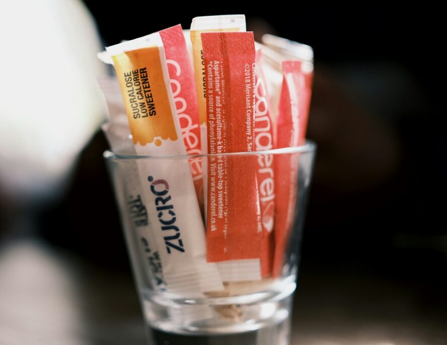 Image of a small glass shot glass full of sweetener packets, including sucralose.
