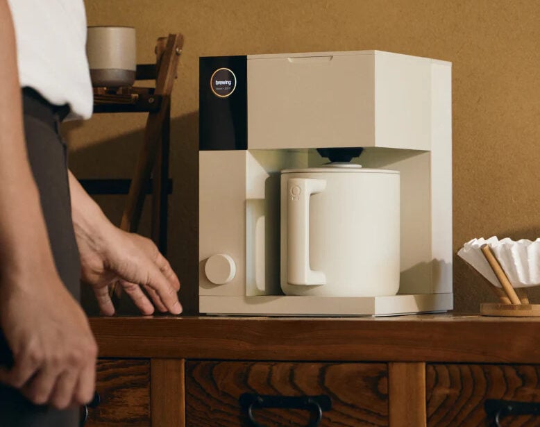 Image of a Fellow Aiden Precision Coffee Maker on a wooden table against a coffee-colored wall.