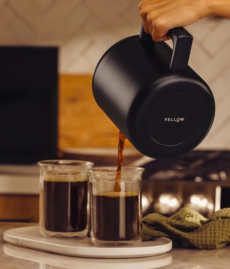 Image of the Fellow Aiden Precision Coffee Maker in matte black, shown with a hand pouring coffee into glass mugs.