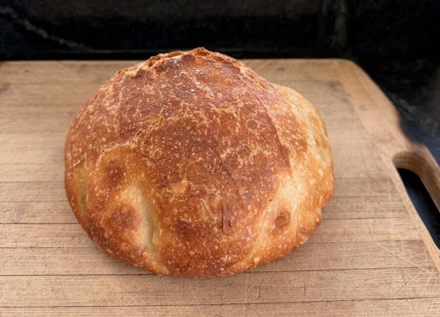 Image of a crusty loaf of no-knead Dutch oven bread shown on a wooden cutting board.