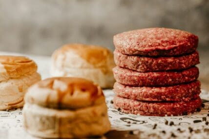 Image of several raw ground beef burgers stacked on top of each other next to buns.