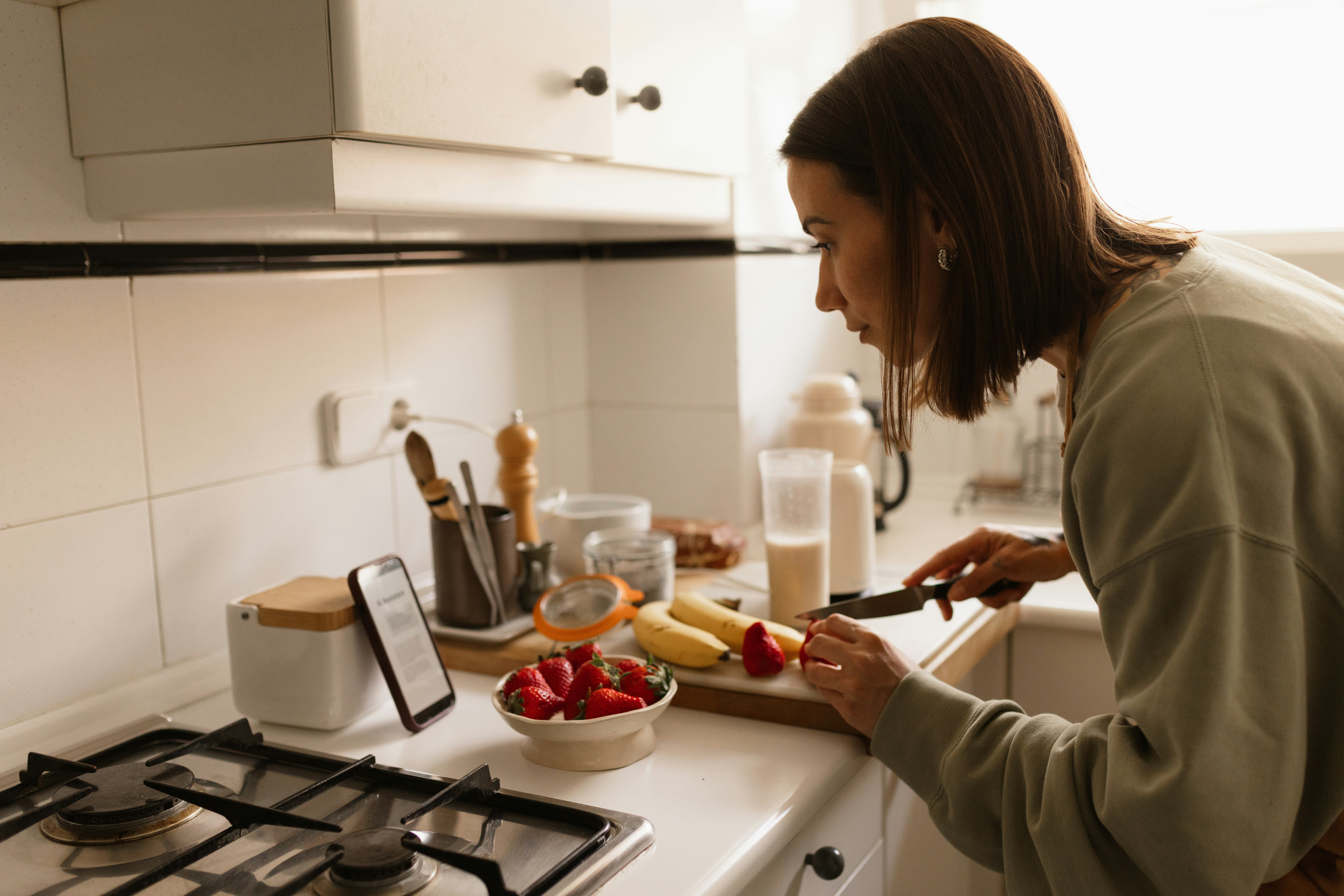 woman cutting up fruit while looking at recipe bent over kitchen counter on her phone.