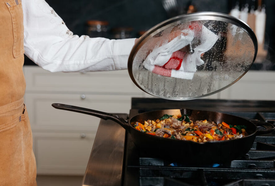 Image of someone at a stovetop lifting a lid off a Made In cast iron skillet being used to sear meat and veggies.